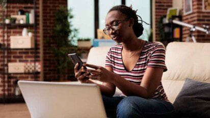 A imagem mostra uma mulher sentada em um sofá em ambiente interno, segurando um celular e olhando atentamente para a tela. Ela usa óculos, camiseta listrada e calça jeans, e está com expressão concentrada. Em primeiro plano, há um notebook branco parcialmente visível, sugerindo que ela está realizando alguma atividade digital. Ao fundo, é possível ver uma estante com objetos decorativos e uma parede de tijolos aparentes, compondo um cenário moderno e acolhedor.