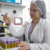 Profissional de laboratório analisando tubo de coleta de sangue em ambiente hospitalar, com bancada organizada, microscópio e diversos tubos identificados ao fundo. Foto: Divulgação/ISGH