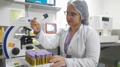 Profissional de laboratório analisando tubo de coleta de sangue em ambiente hospitalar, com bancada organizada, microscópio e diversos tubos identificados ao fundo. Foto: Divulgação/ISGH