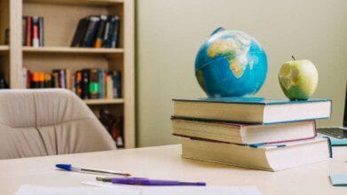 Processos Seletivos 9 Imagem de uma mesa de estudo ou trabalho com três livros empilhados, um globo terrestre e uma maçã verde parcialmente comida sobre os livros. Em primeiro plano, há duas canetas e folhas de papel, sugerindo um ambiente acadêmico. Ao fundo, uma estante com livros variados e uma cadeira de escritório completam a cena, remetendo a um espaço educacional ou de leitura. (Ilha Grande PI)