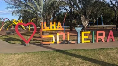 Letreiro colorido com a frase Eu Amo Ilha Solteira em uma praça com árvores e gramado ao fundo sob céu claro.