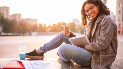 Estudante sentada ao ar livre segurando caderno e caneta, sorrindo, com materiais de estudo e bebida ao lado.