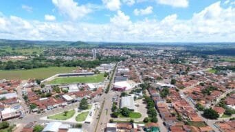 Imagem aérea da cidade de Inhumas-GO, com áreas urbanas, residências, estádio de futebol e vegetação ao fundo em um dia de céu azul com nuvens