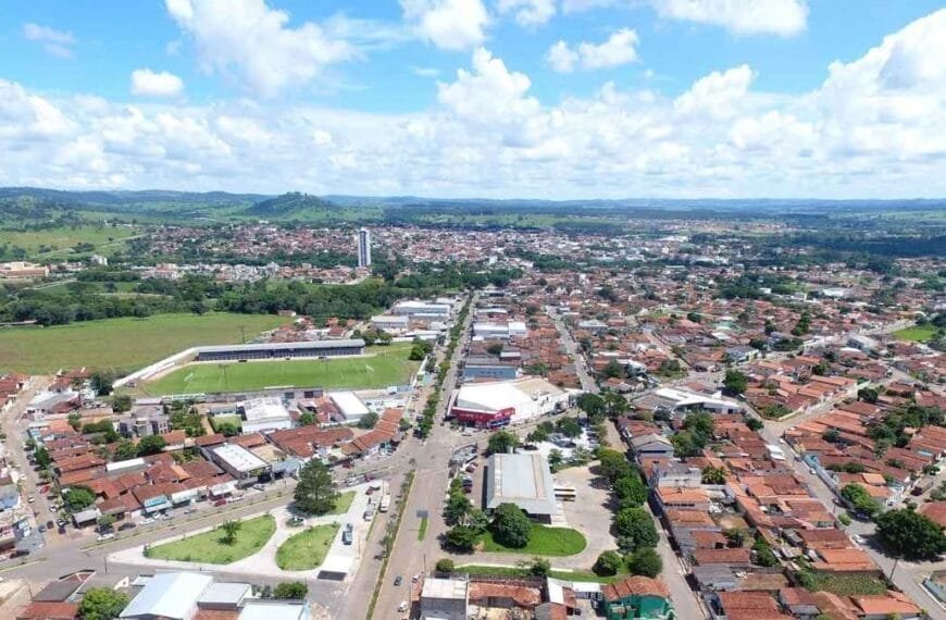 Imagem aérea da cidade de Inhumas-GO, com áreas urbanas, residências, estádio de futebol e vegetação ao fundo em um dia de céu azul com nuvens