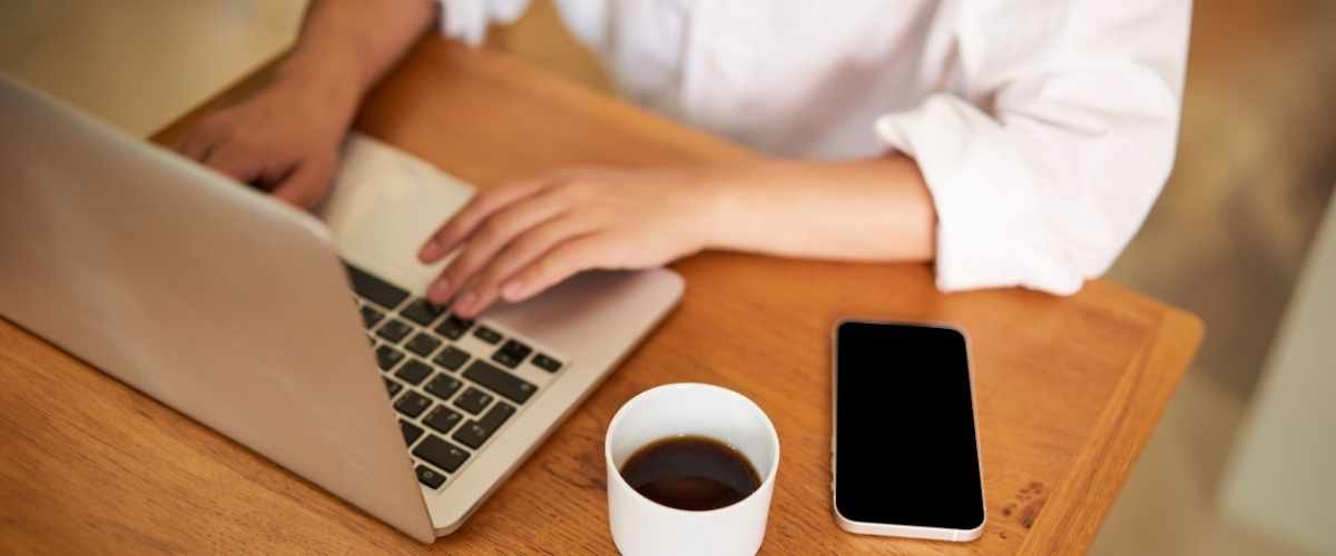Concurso CREMAM: 270 vagas com salários de até R$ 7,2 mil 2 Pessoa utilizando um notebook sobre uma mesa de madeira, com uma xícara de café e um smartphone ao lado. A pessoa veste uma camisa branca de mangas dobradas e está digitando no teclado. A tela do celular está apagada. A cena remete a um ambiente de trabalho ou estudo, transmitindo foco, produtividade e uso de tecnologia para atividades online, como inscrições em concursos.