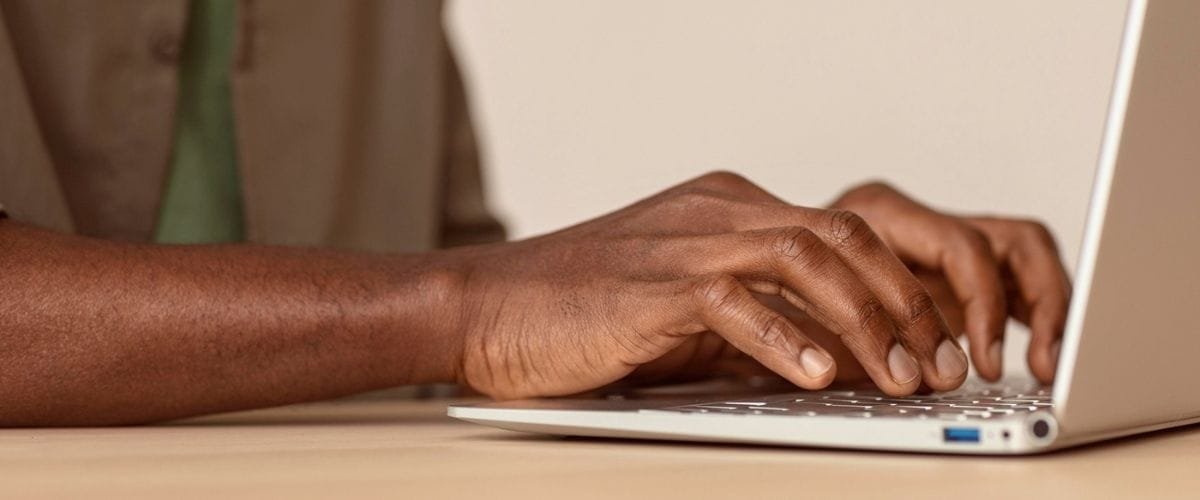 Carmo do Paranaíba MG abre seleção com salário de R$ 3 mil 2 Pessoa digitando em um notebook branco sobre uma mesa clara. A imagem mostra apenas os braços e as mãos de um homem com pele escura, vestindo camisa bege. Ele está focado na digitação, sugerindo atividade como inscrição online ou preenchimento de formulário. O ambiente é neutro e bem iluminado, com destaque para a ação de uso do computador.