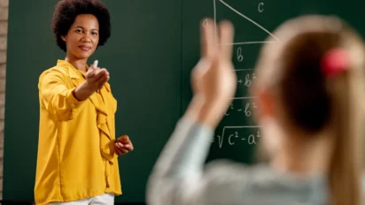 Professora em sala de aula aponta para aluna que levanta a mão, com equações matemáticas escritas no quadro. Foto: Drazen Zigic/Freepik