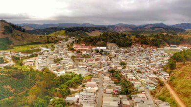 Processos Seletivos 5 Vista aérea de Irupi-ES com casas, ruas e áreas verdes cercadas por morros e vegetação sob céu nublado. Foto: Irupiense/Wikipédia