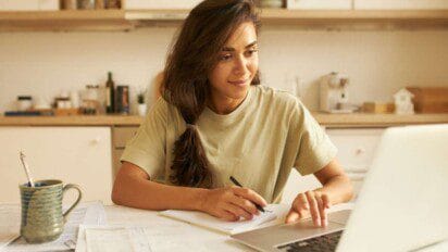 Itaitinga - Mulher jovem com cabelos castanhos e amarrados em um rabo de cavalo estuda em casa, sentada à mesa com notebook aberto, caderno e caneta na mão. Ela sorri levemente enquanto observa a tela do computador, demonstrando concentração. Ao lado, há uma caneca com bebida quente e alguns papéis espalhados. O ambiente é uma cozinha organizada e iluminada, sugerindo um clima de tranquilidade e produtividade.