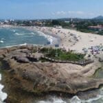 Vista aérea da orla de Itapoá-SC com praia movimentada, mar, estrutura de deck sobre rochas e cidade ao fundo.