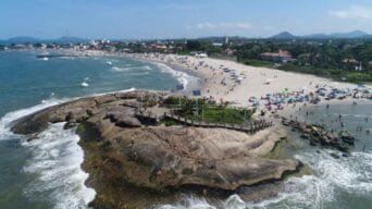 Vista aérea da orla de Itapoá-SC com praia movimentada, mar, estrutura de deck sobre rochas e cidade ao fundo.