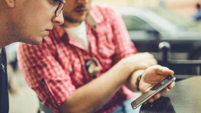 Dois homens sentados observam a tela de um celular com atenção, em ambiente externo próximo a carros estacionados.