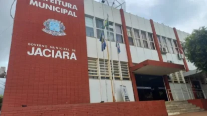Fachada da Prefeitura de Jaciara com paredes vermelhas e brancas, brasão do município, janelas amplas e mastros com bandeiras em frente ao prédio. Foto: Divulgação/Prefeitura de Jaciara-MT