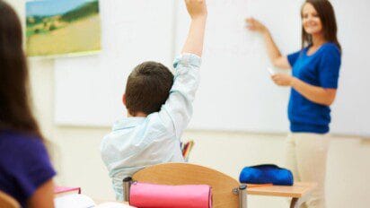 Sala de aula com ambiente iluminado, onde uma professora está em pé à frente de um quadro branco, explicando algum conteúdo para os alunos. Ela sorri e segura um marcador, enquanto aponta para o quadro. Em primeiro plano, um menino com camisa clara está sentado de costas e levanta o braço, aparentemente para fazer uma pergunta. Outras crianças também estão presentes, sentadas em carteiras escolares com estojos e cadernos sobre a mesa. A imagem transmite um ambiente educativo e participativo. (Jaguaretama )