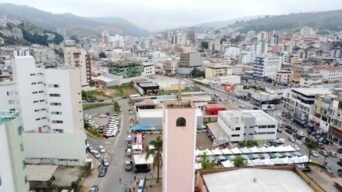 Vista aérea de João Monlevade-MG destaca o centro urbano com prédios, tráfego intenso, feira e torre de relógio, cercados por morros e clima nublado.