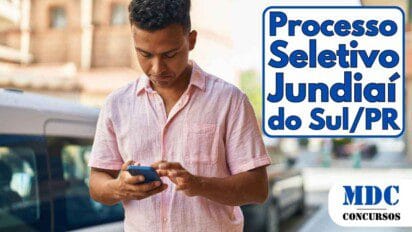 Homem jovem de camisa rosa olhando para o celular enquanto está na calçada, com carros estacionados e prédios ao fundo em um dia ensolarado - Jundiaí do Sul oferece salários de até R$ 3 mil em Processo Seletivo