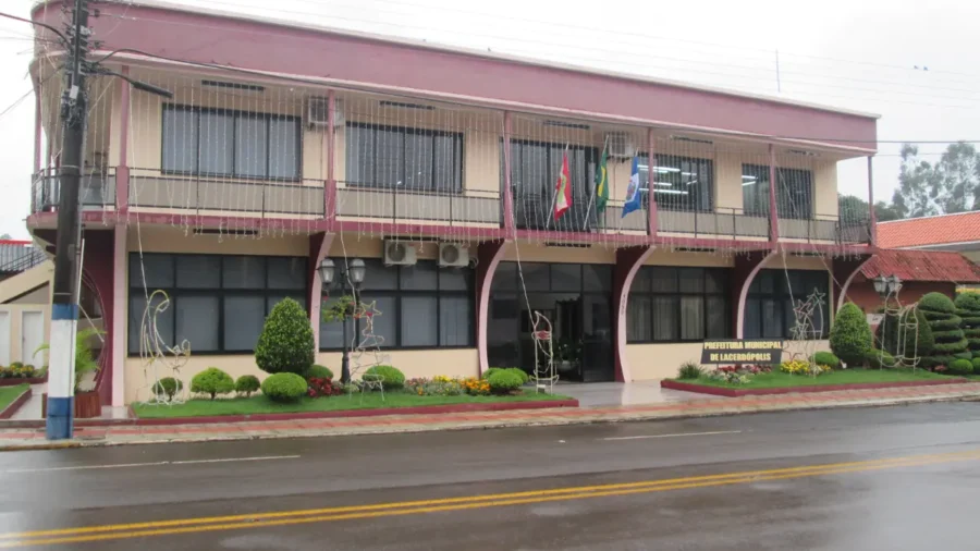 Fachada frontal da Prefeitura Municipal de Lacerdópolis em Santa Catarina, um prédio de dois pavimentos em tons de bege e bordô com bandeiras hasteadas na sacada e decorações natalinas no jardim frontal sob dia chuvoso.