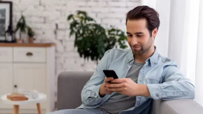 Homem sentado em um sofá usando o celular em um ambiente doméstico iluminado. Foto: Freepik