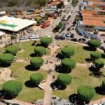 Vista aérea de uma praça circular com árvores podadas simetricamente, caminhos pavimentados e pessoas circulando; ao fundo, tendas brancas e casas com telhados alaranjados