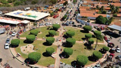 Processos Seletivos 8 Vista aérea de uma praça circular com árvores podadas simetricamente, caminhos pavimentados e pessoas circulando; ao fundo, tendas brancas e casas com telhados alaranjados