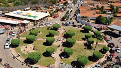 Vista aérea de uma praça circular com árvores podadas simetricamente, caminhos pavimentados e pessoas circulando; ao fundo, tendas brancas e casas com telhados alaranjados