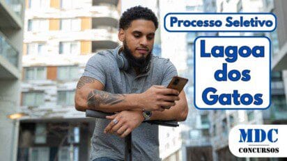 Homem de barba e cabelos cacheados, usando camisa polo cinza e fones de ouvido no pescoço, apoia-se em um patinete elétrico enquanto olha para o celular em um ambiente urbano com prédios modernos ao fundo - Lagoa dos Gatos (PE) Abre Vagas com Salário de R$ 2.824 para ACS em Processo Seletivo