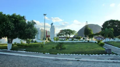 Praça arborizada em Lajedão-BA com gramado, árvores, calçada de pedras e prédio branco moderno ao fundo, tendo morro rochoso ao horizonte. Foto: Vilamir Azevedo/Wikipédia