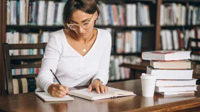 Mulher estudando em biblioteca, escrevendo em caderno com livros e café sobre a mesa. Foto: senivpetro/Freepik