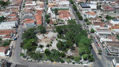 Processos Seletivos 5 Vista aérea do centro de Limoeiro-PE, destacando uma praça arborizada cercada por ruas, casas e comércios.