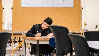 Jovem sentado em sala de provas, escrevendo em uma folha sobre a mesa, com cadeiras e divisórias ao fundo.