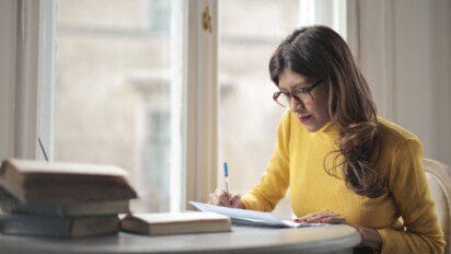Mulher de óculos com blusa amarela escrevendo em papel sobre uma mesa, ao lado de livros, em frente a uma janela iluminada. Foto: andreas/Freepik