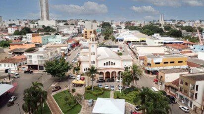 Vista aérea do centro de Luziânia-GO com destaque para uma igreja de fachada clara e torre com cúpula dourada, rodeada por ruas, carros e prédios comerciais.