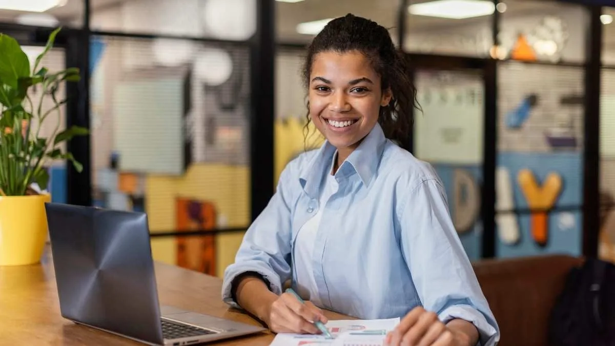 Mulher sorridente sentada à mesa com notebook e documentos, em ambiente de escritório moderno e colorido.