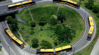 Processos Seletivos 3 Ônibus articulados amarelos da MOBI Rio circulando em uma rotatória cercada por área verde.