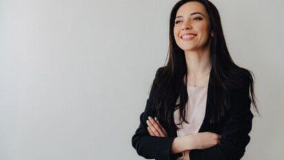 Mulher sorridente de braços cruzados usando blazer preto e blusa clara em fundo branco. Foto: pressahotkey/Freepik