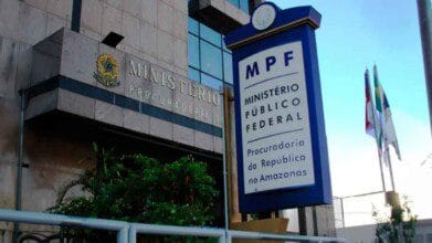 Processos Seletivos 5 Fachada da sede do Ministério Público Federal no Amazonas, com destaque para a placa institucional azul do MPF indicando a Procuradoria da República no estado. O edifício apresenta arquitetura moderna com revestimento em granito e vidro espelhado. À direita da imagem, aparecem mastros com bandeiras hasteadas, simbolizando o Brasil, o Amazonas e outras entidades. A vegetação na base do prédio complementa o cenário urbano oficial.