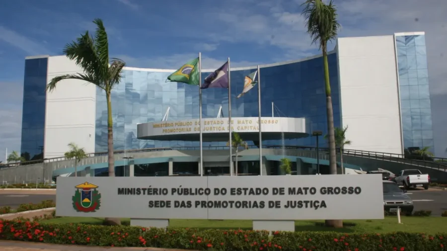 Fachada da sede das Promotorias de Justiça do Ministério Público do Estado de Mato Grosso em Cuiabá (MPMT), exibindo um edifício moderno espelhado com bandeiras e palmeiras à frente.
