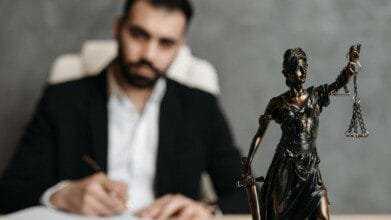 Estátua da deusa da justiça em primeiro plano com balança e espada, e um homem desfocado ao fundo escrevendo em um documento.