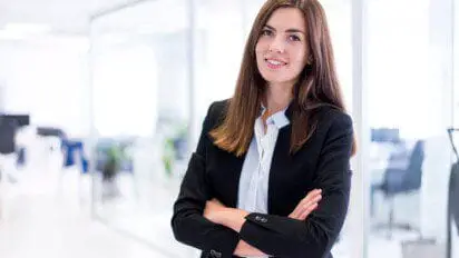 Mulher jovem de terno preto e camisa branca sorrindo com os braços cruzados em um ambiente corporativo iluminado.