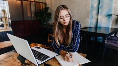 Mulher com óculos escreve em caderno enquanto estuda com notebook em mesa de madeira. Foto: lookstudio/Freepik