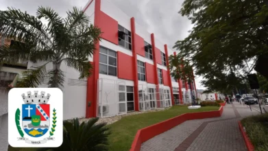 Maricá/RJ abre processo seletivo com 34 vagas e salários de até R$ 5 mil 4 Prédio público de Maricá com fachada branca e vermelha, janelas amplas, área ajardinada, calçada lateral e brasão do município no canto da imagem. Foto: Montagem MDC Concursos/Divulgação/Prefeitura de Maricá-RJ