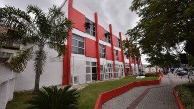 Prefeitura Municipal de Maricá, no Rio de Janeiro, um prédio moderno de três andares com fachada vermelha e branca, cercado por gramado e algumas palmeiras sob um céu nublado