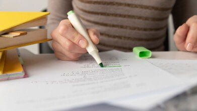 Processos Seletivos 2 Pessoa sublinhando um texto com marcador verde em uma folha de prova, ao lado de livros e materiais de estudo.