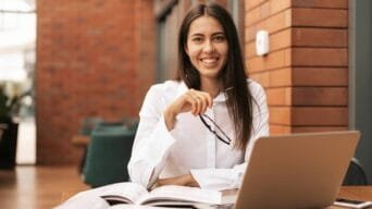 Mulher sorridente com camisa branca sentada à mesa com laptop, livro aberto e segurando óculos, em ambiente moderno com parede de tijolos.