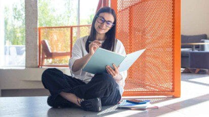 Mulher jovem de cabelos longos e óculos sentada no chão de um ambiente moderno e iluminado, sorrindo enquanto escreve em um caderno ou apostila. Ela está cercada por materiais de estudo, como livros e cadernos, sugerindo um momento de preparação para concurso público ou prova. Ao fundo, há móveis contemporâneos e uma divisória laranja vazada, indicando um espaço acolhedor e funcional. (Mondaí)