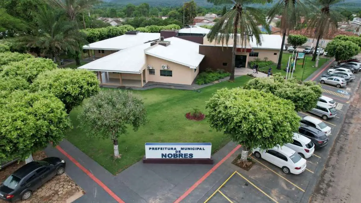 Vista aérea da sede da Prefeitura Municipal de Nobres, cercada por áreas verdes, árvores e estacionamento com diversos carros.