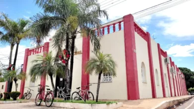 Fachada da Prefeitura de Nossa Senhora do Livramento com prédio histórico em tons de bege e vermelho, palmeiras na calçada, bicicletas estacionadas e céu azul ao fundo. Foto: Divulgação/Prefeitura de Nossa Senhora do Livramento-MT