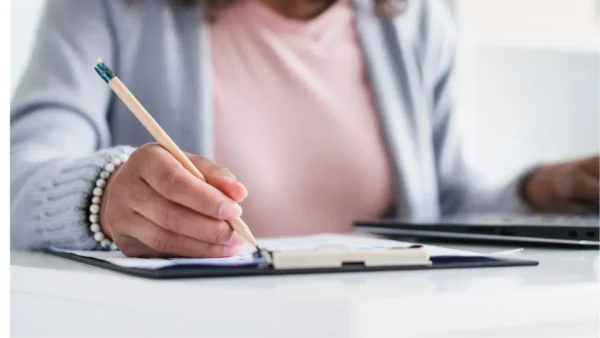 Nova Floresta/PB abre concurso com 136 vagas e salários de até R$ 14 mil 5 Pessoa escrevendo em um caderno com lápis sobre uma mesa, com notebook ao lado, representando estudo ou preenchimento de documentos. Foto: Freepik