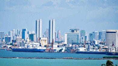 Processos Seletivos 8 Vista do porto de Recife com navios atracados em primeiro plano e prédios altos ao fundo sob céu azul.