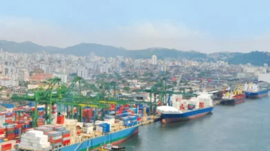 Processos Seletivos 6 Vista aérea do Porto de Santos com navios cargueiros atracados, contêineres coloridos e guindastes, tendo a cidade e morros ao fundo. Foto: Divulgação/Prefeitura de Santos-SP